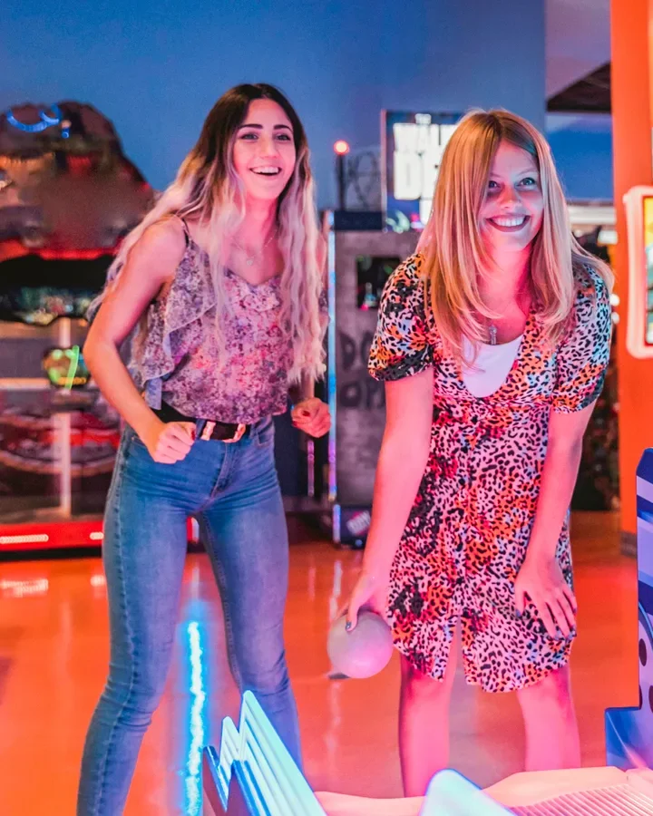 Two friends laughing over a skee-ball lane in a neon-lit LA arcade