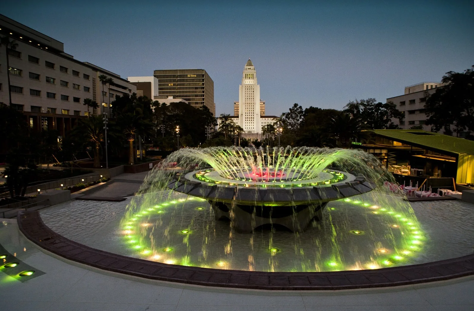 Round fountain lit green at dusk with a civic tower behind