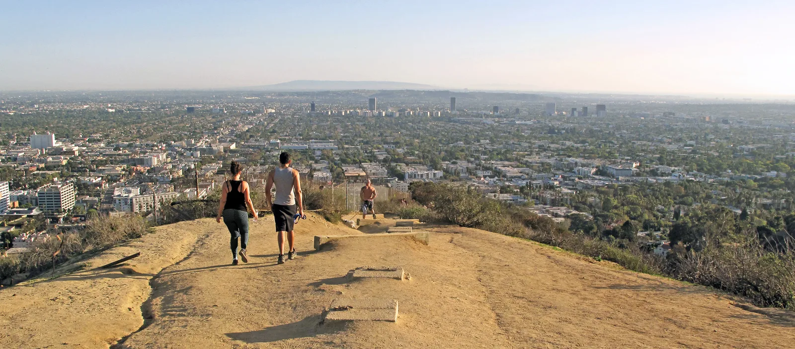 Two hikers on a dirt ridge trail above a hazy city