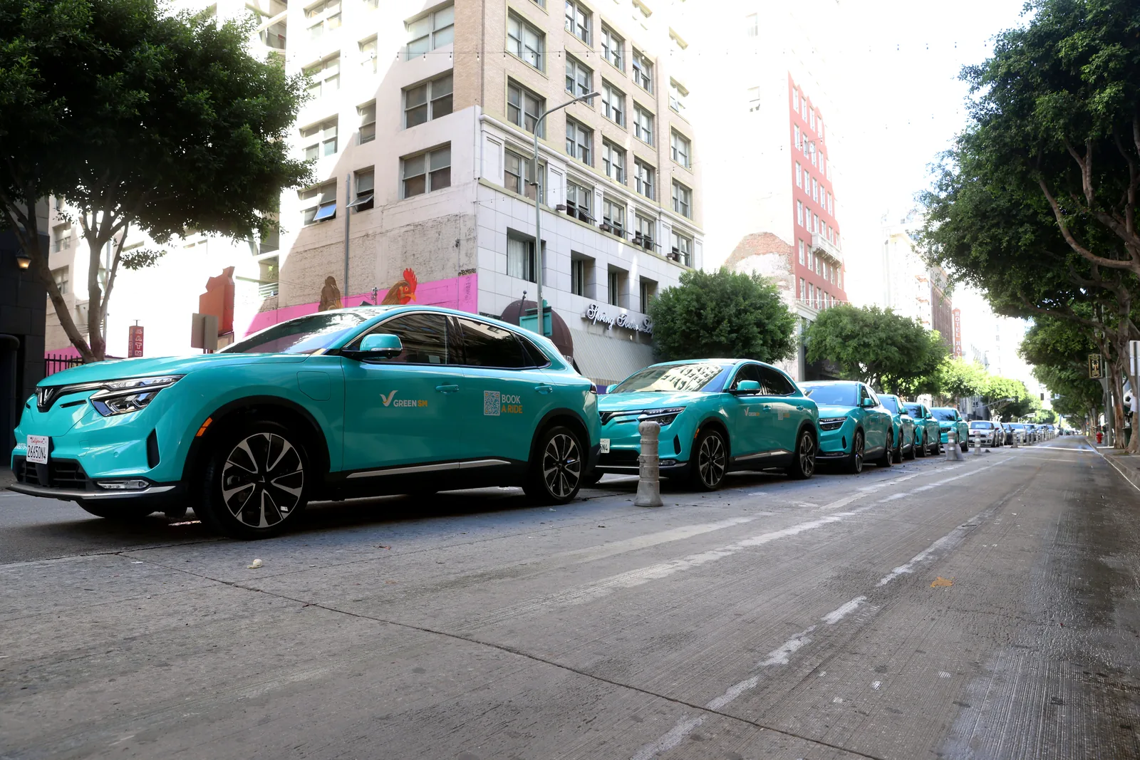 A row of cyan Green SM VinFast VF 8 SUVs parked along a downtown Los Angeles street