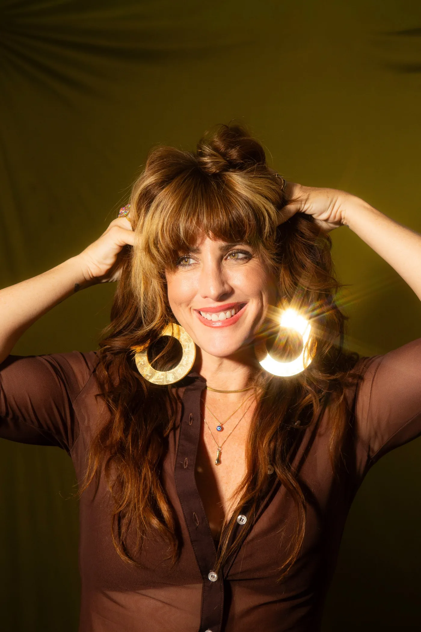 Jennifer Sodini smiling with her hands in her hair, wearing gold zodiac hoop earrings against an olive backdrop
