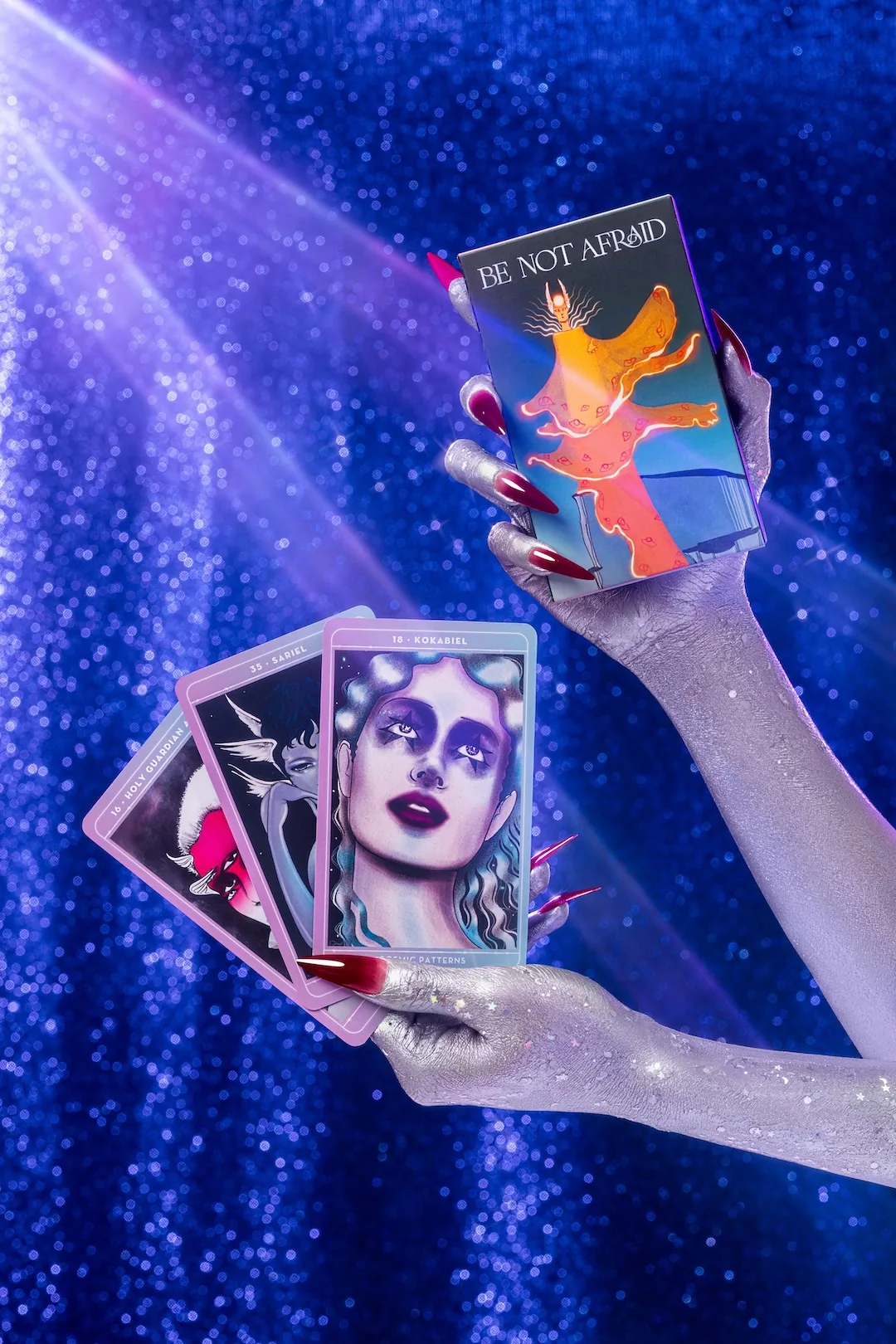 Silver-painted hands holding the Be Not Afraid oracle deck box and three fanned angel cards against a blue sequin backdrop