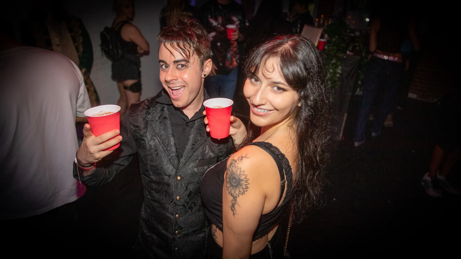 Two guests raise red cups and smile in a dark room