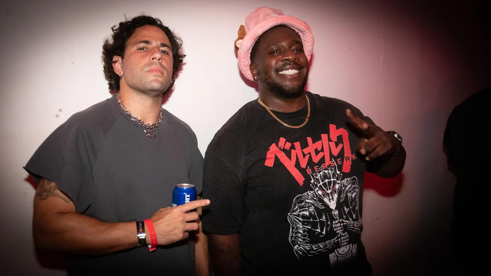 Two guests pose against a pink-lit wall, one holding a canned drink