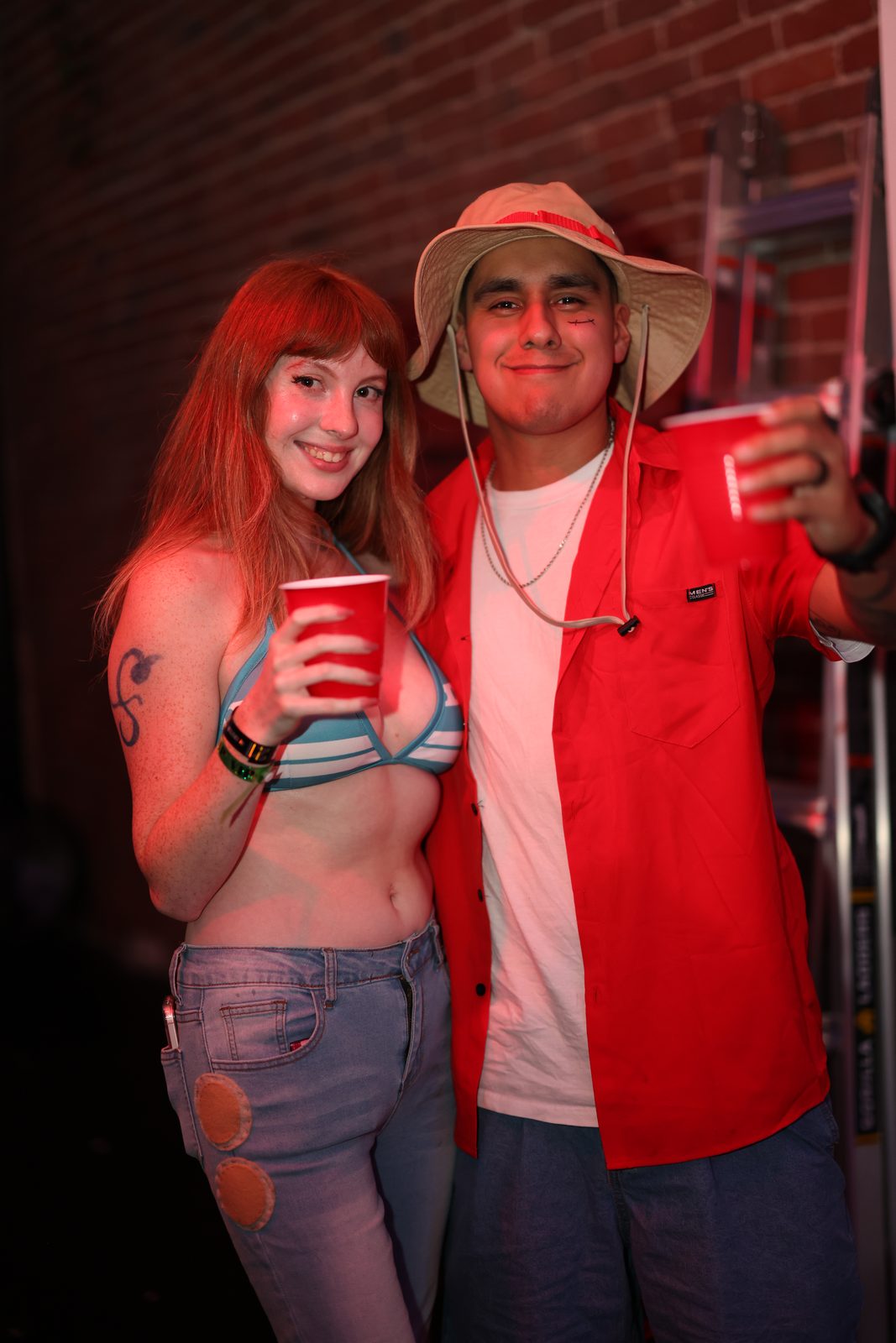 Two guests hold red cups against a brick wall, one in a sun hat