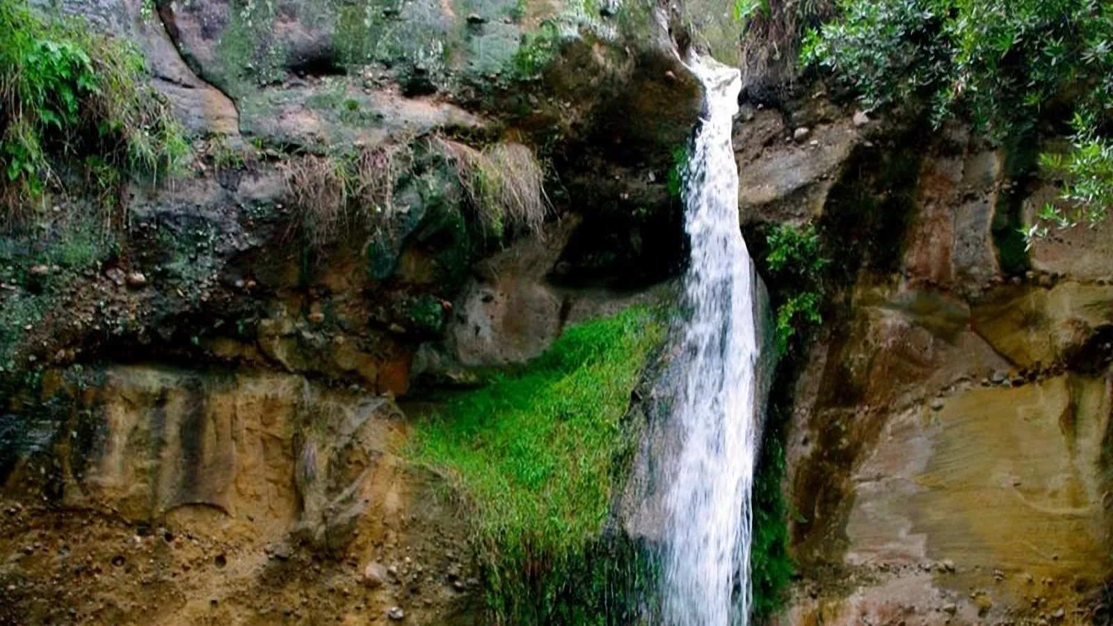 Narrow waterfall dropping over mossy sandstone cliffs in a shaded canyon
