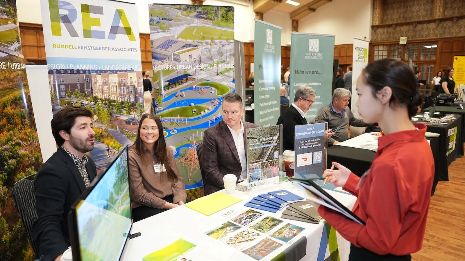 Three exhibitors behind a booth table talk with a visitor holding brochures