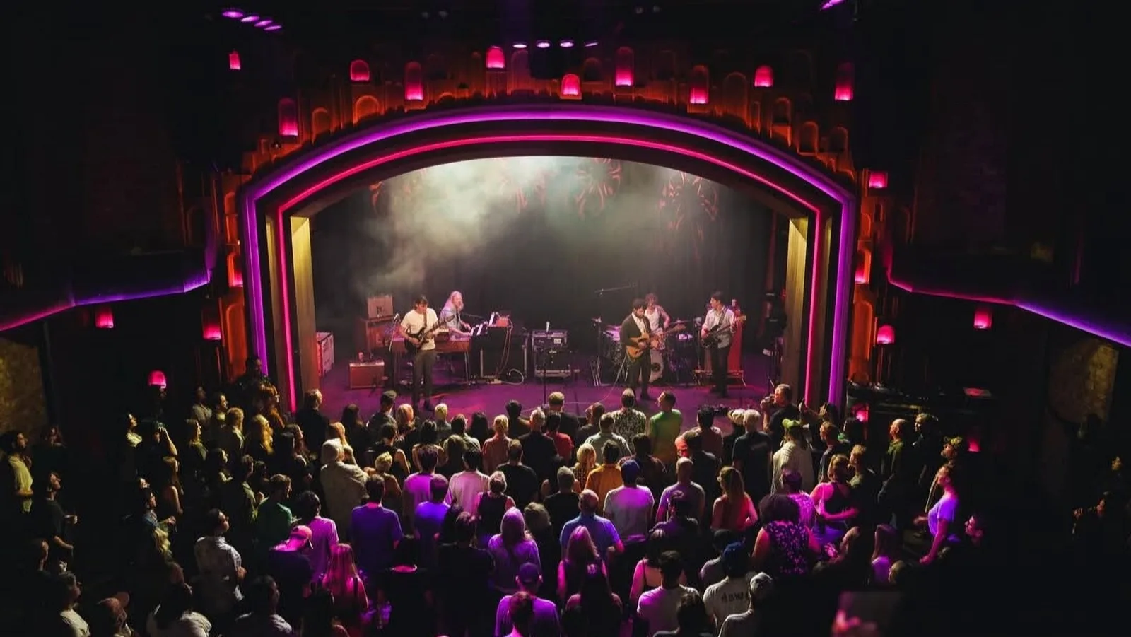 Band performing on a theatre stage above a packed pink lit audience