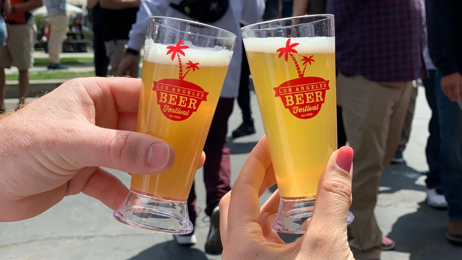 Two hands clinking pint glasses branded Los Angeles Beer Festival outdoors