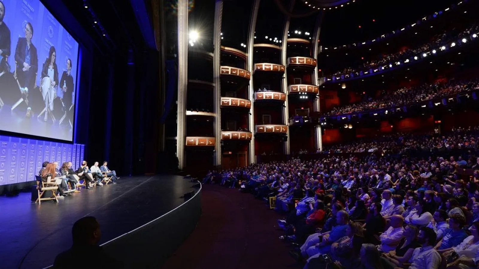 Packed concert hall with tiered balconies and speakers seated on the stage