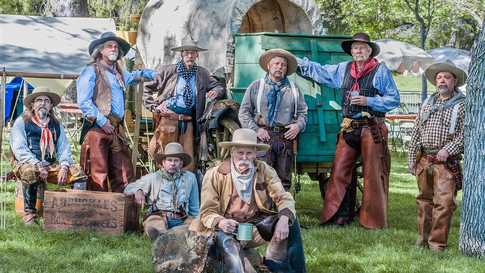 Six cowboy reenactors pose in chaps beside a green chuckwagon on grass