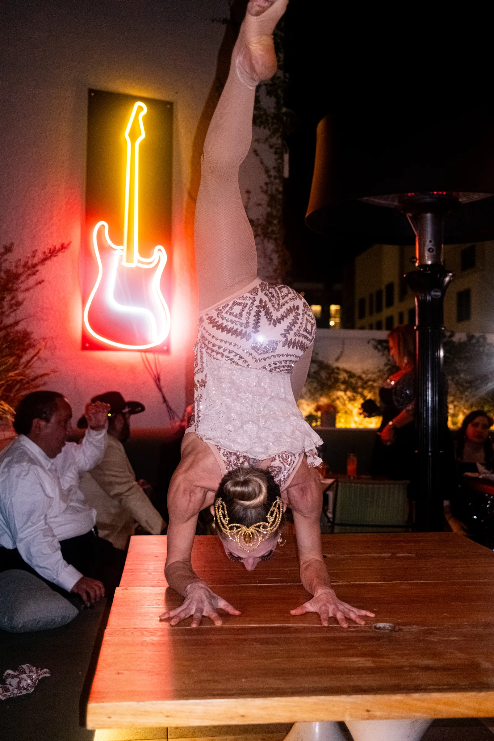 A performer does a handstand beside a neon guitar sign on the patio