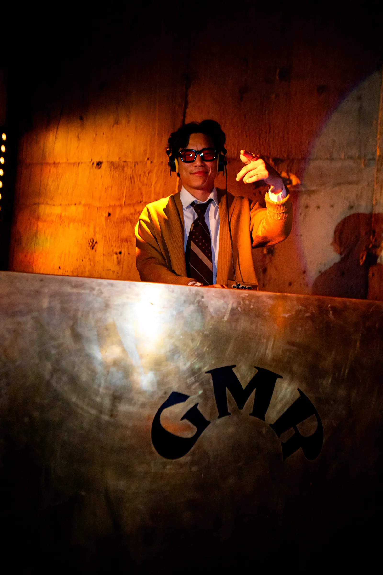 A DJ in an orange suit and headphones stands behind a metal booth