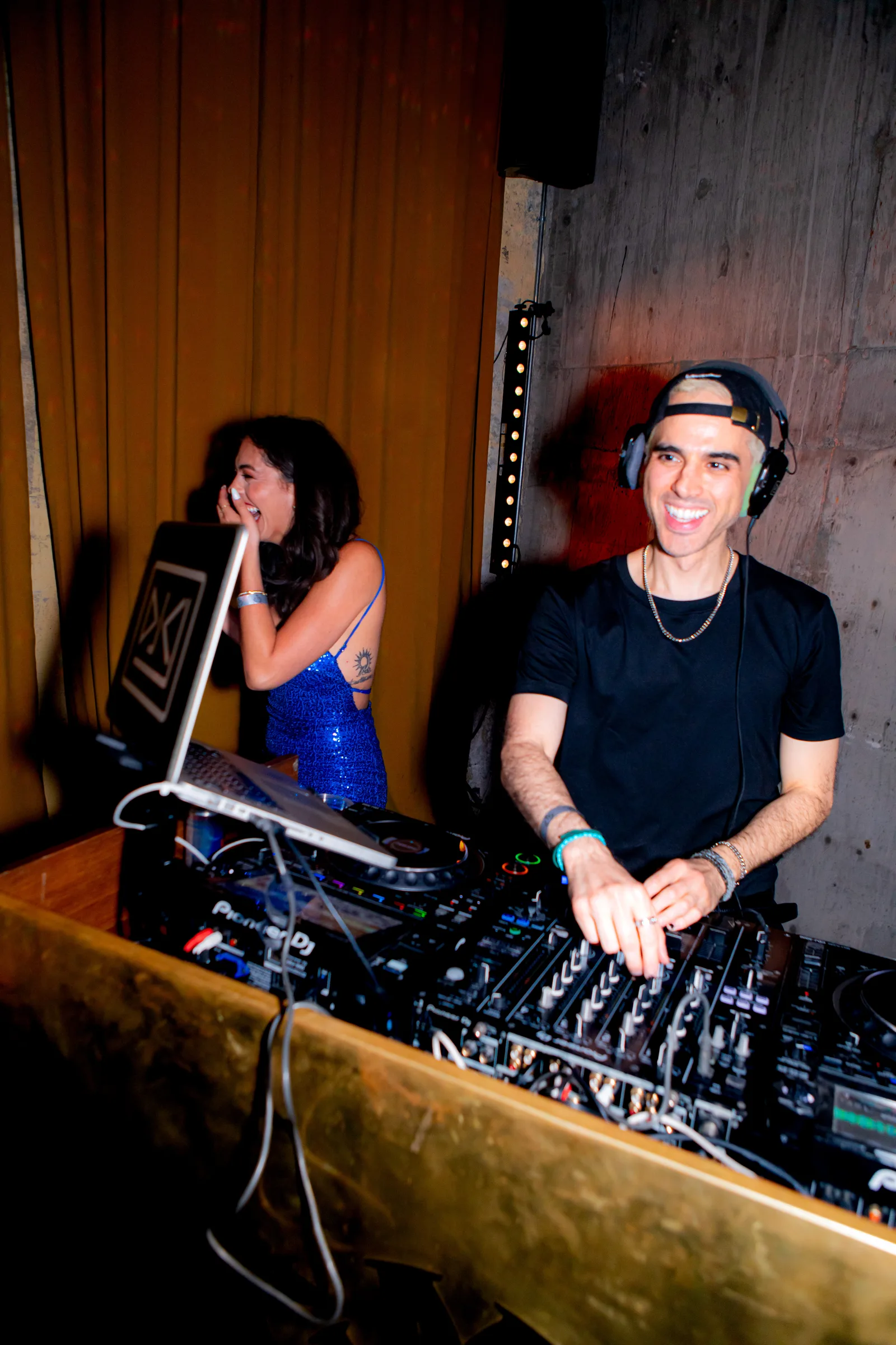 A DJ in a backwards cap mixes while a guest looks on