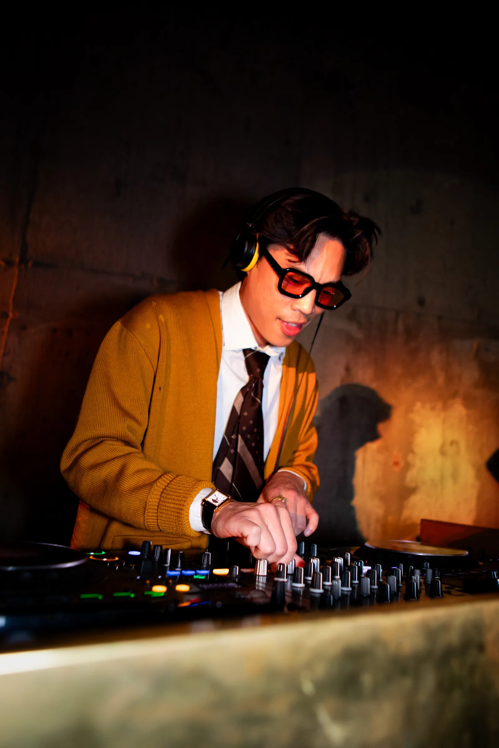 A DJ in a mustard cardigan and dark sunglasses cues a track