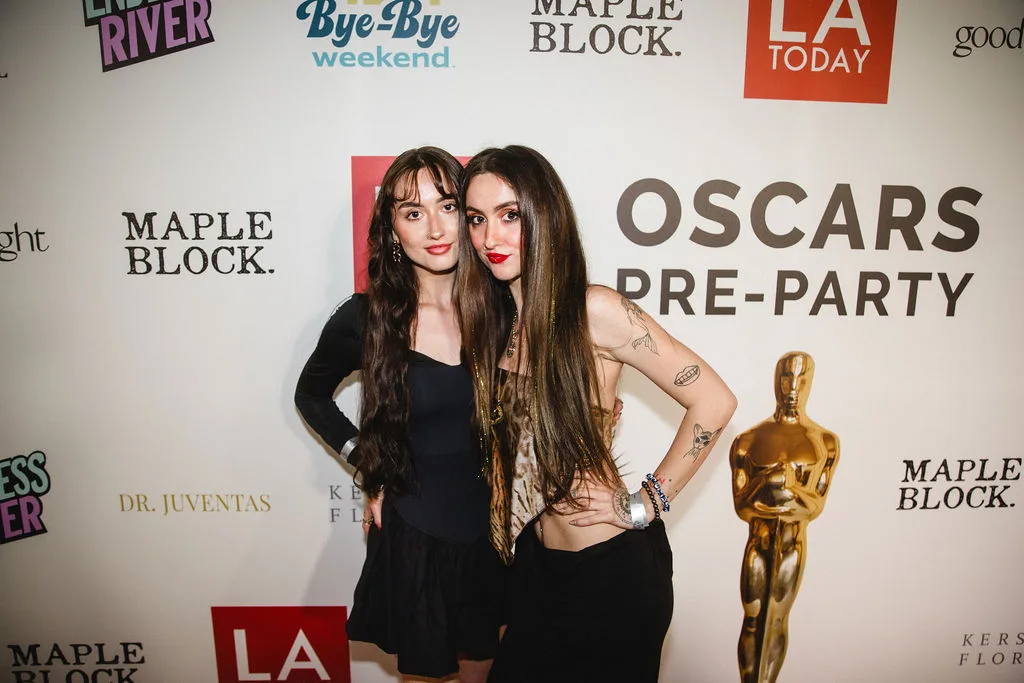 Two guests in black pose beside the gold statuette at the banner