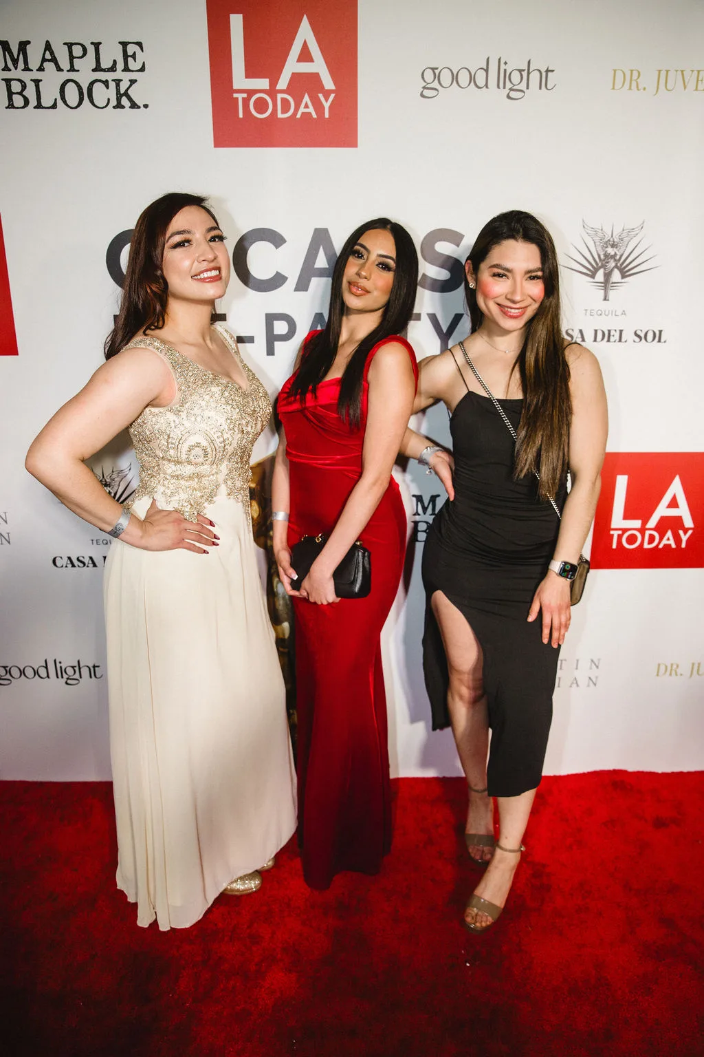 Three guests in gowns pose together against the sponsor backdrop