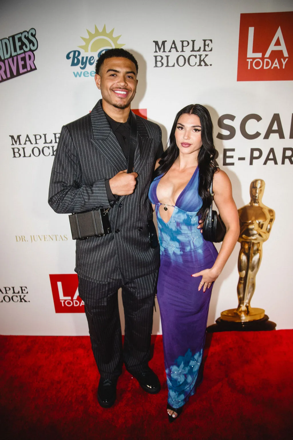 Two guests pose beside a gold statuette at the pre-party backdrop