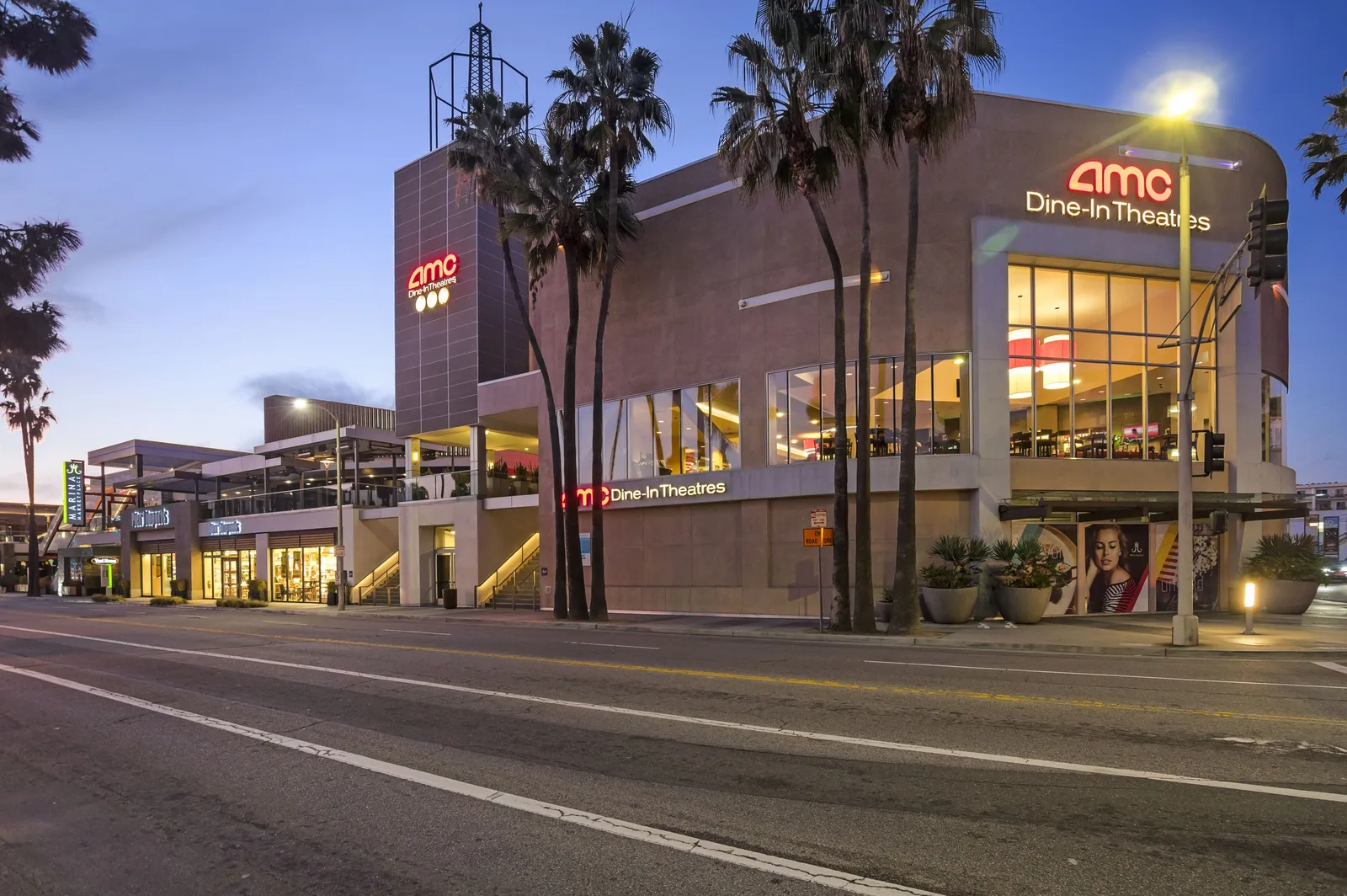 AMC Dine In Theatres building lit at dusk with palms and an empty road