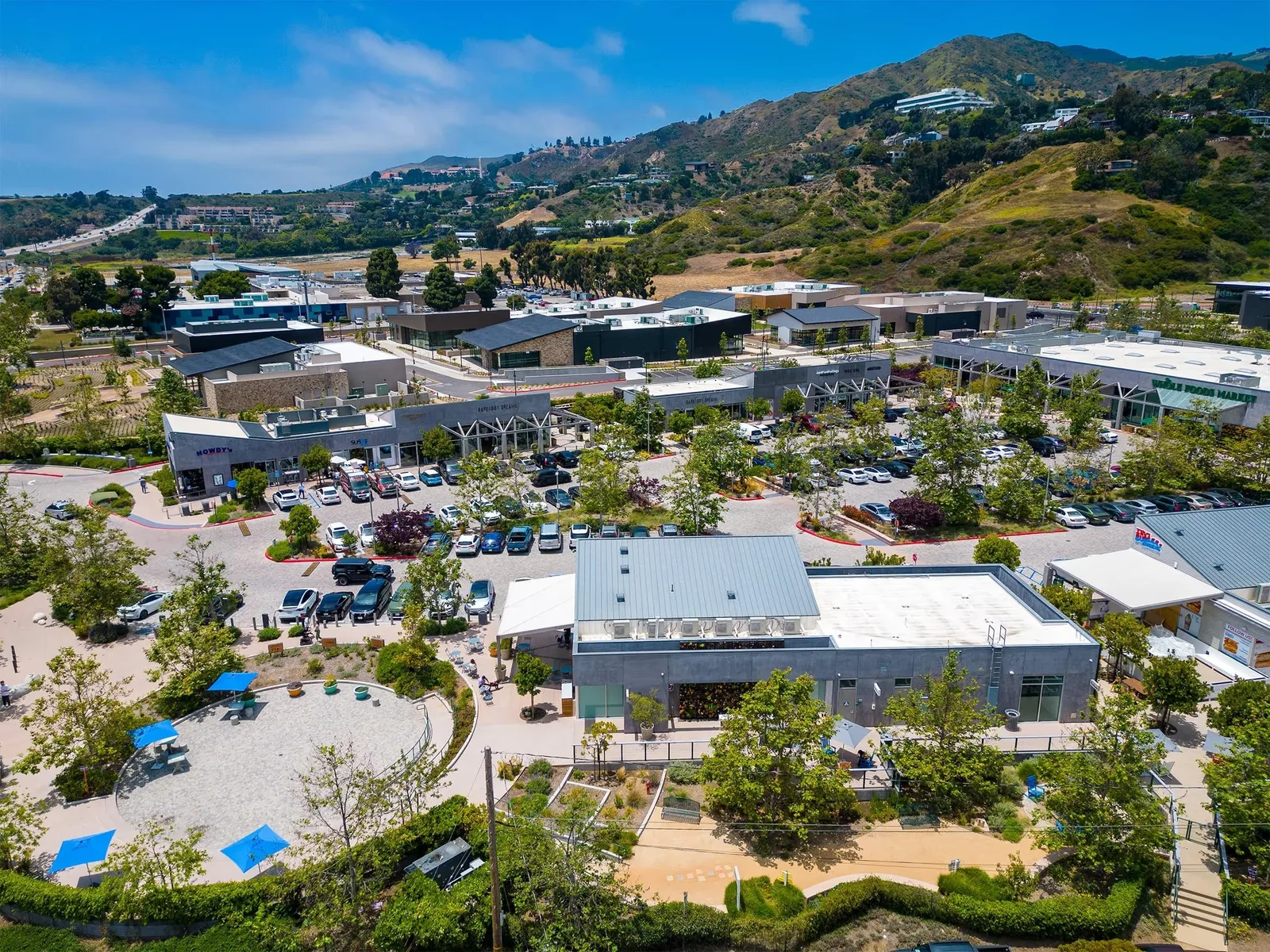 Aerial of low rise shops and parking backed by green hills