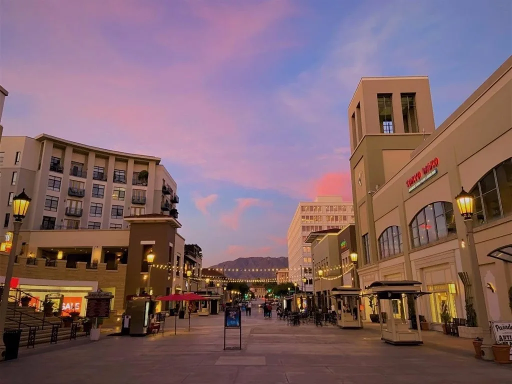 Outdoor shopping street at dusk, warm lit storefronts under a pink sky