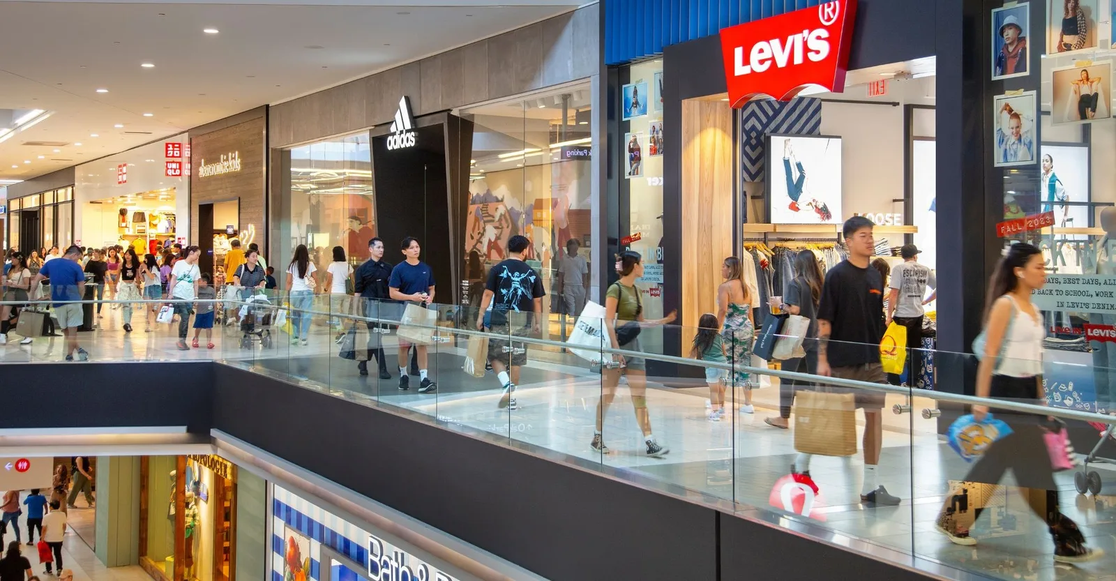 Shoppers pass Levi's and Adidas storefronts along a glass railed mall balcony
