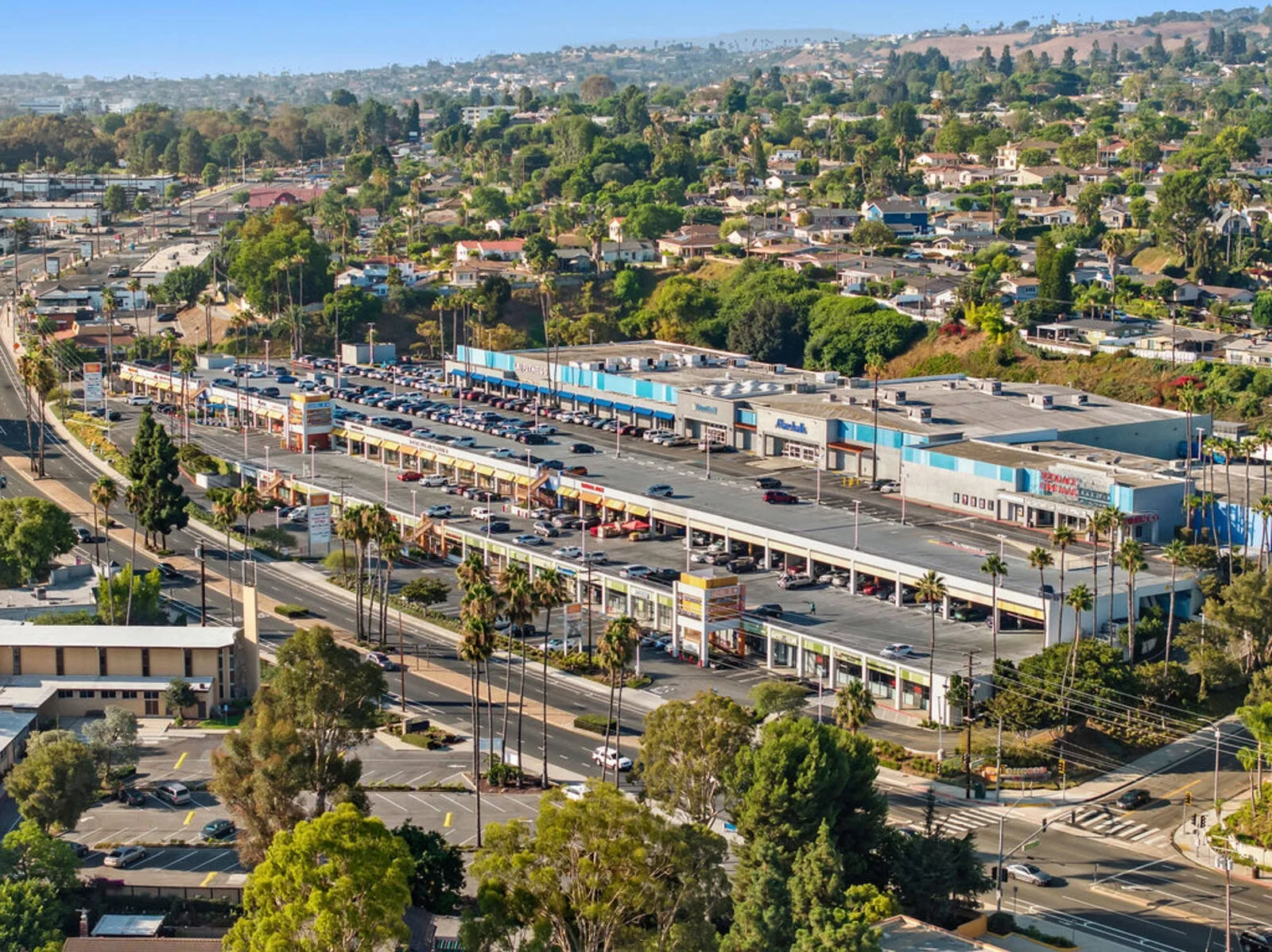 Aerial of long strip mall roofs and parking with hillside housing beyond