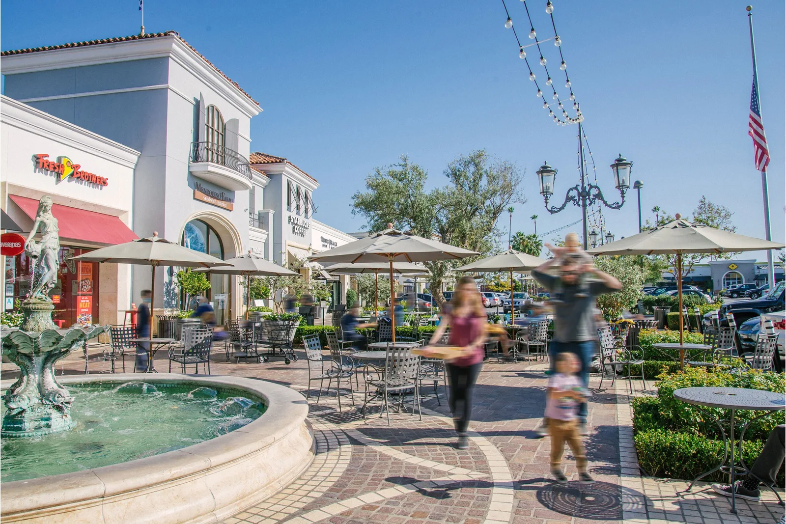 Circular fountain and umbrella tables in a sunny plaza with visitors walking