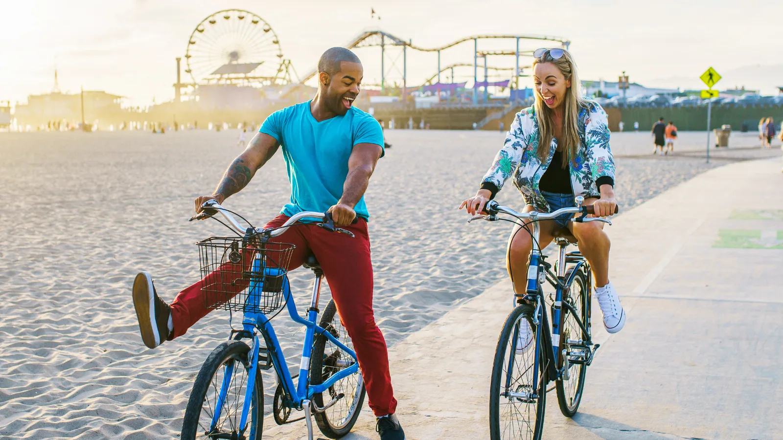 A couple biking on the Santa Monica boardwalk with the Santa Monica Pier in the background.