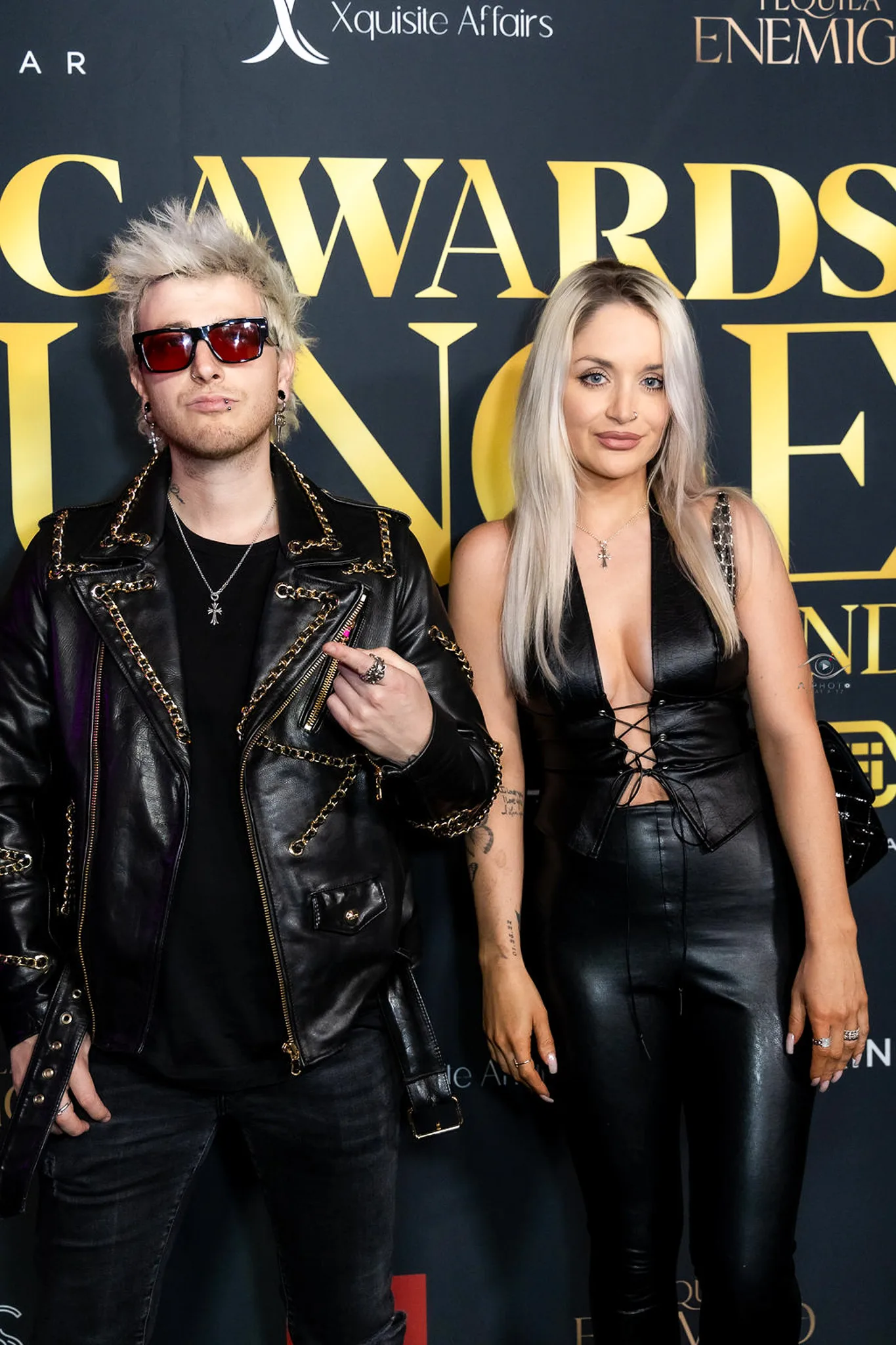 Two guests in black leather looks pose at the gold sponsor backdrop