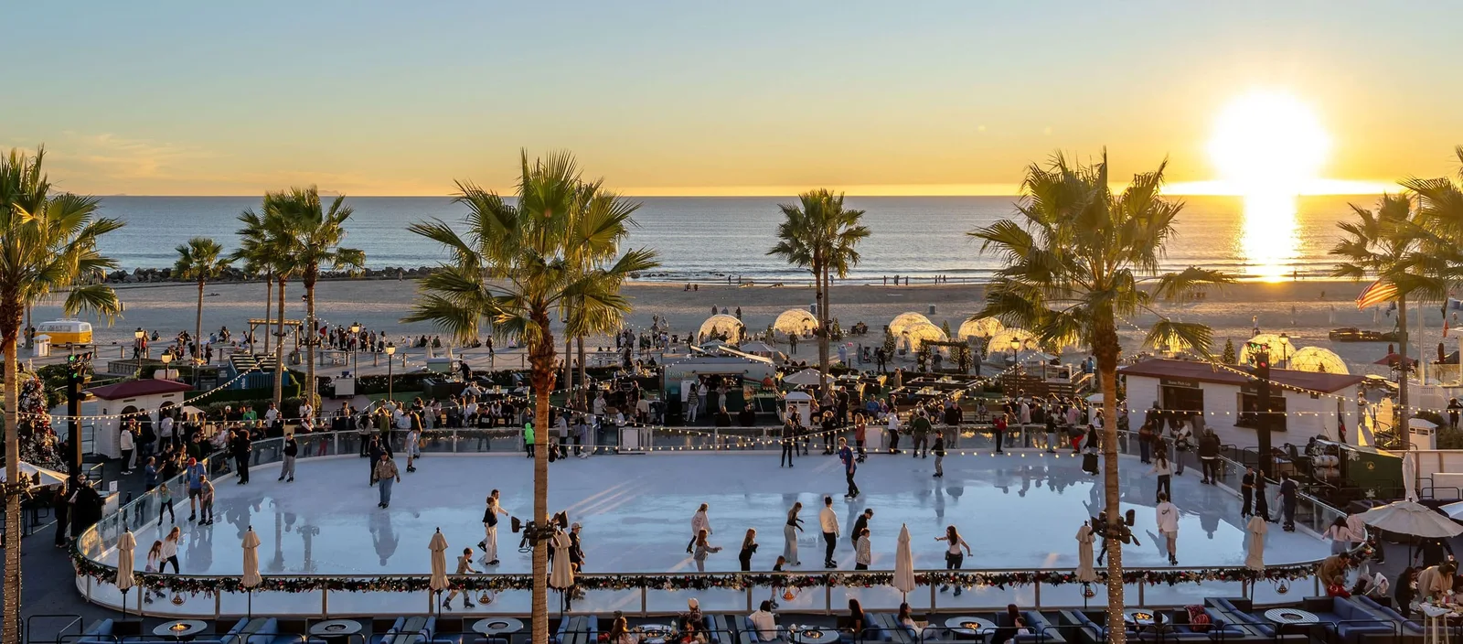 Skaters circle an outdoor rink beside the beach, palms lit at sunset