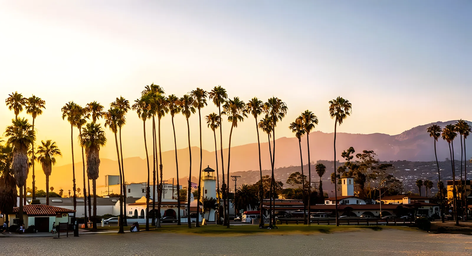Tall palms silhouetted against a golden sunset over a low coastal town and hills